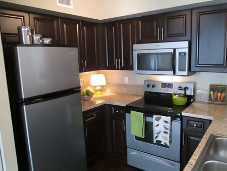 Enclosed kitchen in model apartment home with stainless steel refrigerator, microwave, stove/oven, and double sinks. Granite countertops with plentiful storage in dark wood cabinetry