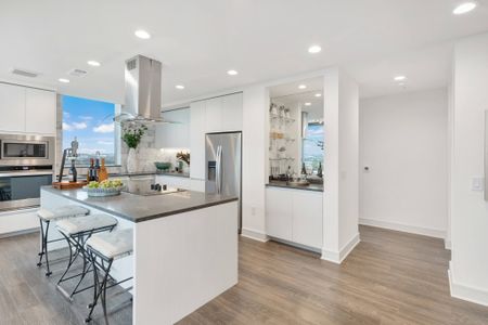 Modern kitchen with stainless steel appliances and overhead range hood, minimalist white handleless cabinets, wood-style floors, and a dry bar.