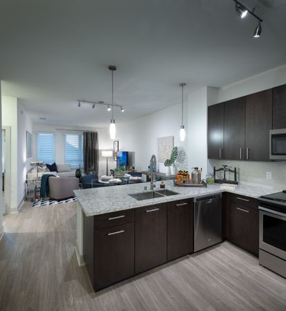 Open-concept kitchen, dining, and living space with ash-gray wood-style floors, dark modern wood cabinets, stainless steel appliances, granite countertops, and a spacious living area with windows and a glass door leading to the balcony.