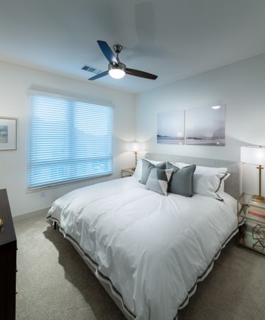 Bedroom with a king-sized bed, two side tables, a dresser, a large window, white walls, a ceiling fan, and beige carpeting.