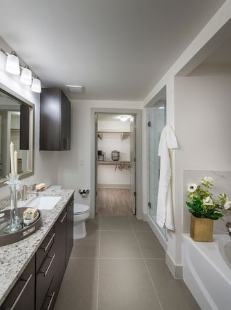 Bathroom with a tub, frameless glass walk-in shower, gray floor tiles, and an attached walk-in closet with wood-style floors.