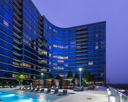 Elevated pool deck with three cabanas and 10 in-pool lounge chairs in front of the backdrop of the upper floors of Buckhead Village, which curve around the pool deck and reflect the dark blue dusk sky.