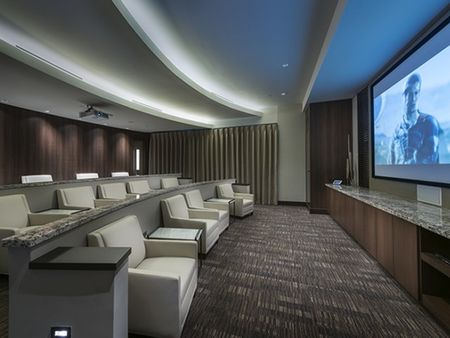 Theater with three elevated rows of large, leather seats and side tables facing a large projector screen. The walls are covered in dark wood panels and there are blackout curtains over the window.