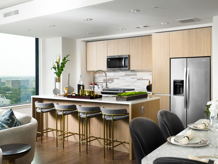 Open-concept kitchen with stainless-steel appliances, modern light wood cabinetry, an island with four bar stools, dark wood-style floors and a city view through the large window.