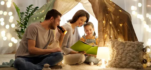 man holding flashlight to book with woman and child in indoor blanket fort