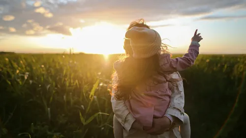 Back view of a person holding a child wearing a beanie, standing in a lush green field at sunset