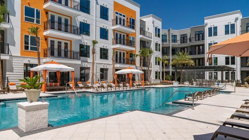 Modern apartment complex with a resort-style pool, sun loungers, and cabanas under a clear blue sky.
