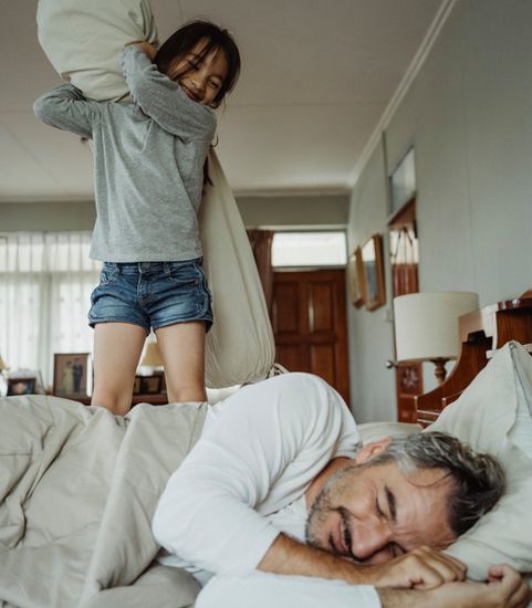 young girl prepping to playfully hit pillow against older man lying down on bed