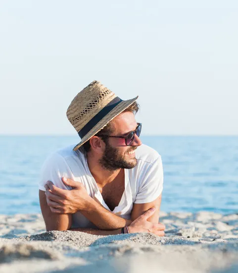 man in hat laying on sandy beach in front of water in daytime