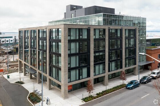 Modern five-story building with large windows, brick facade, and a rooftop area. Several cars are parked on the adjacent street.