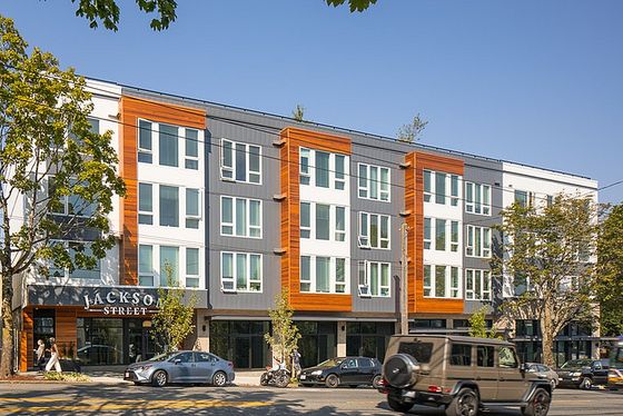 A modern, four-story apartment building with a contemporary design, featuring large windows, dark gray and white paneling, and warm wood accents. The building has a sign reading 'JACKSON STREET' on the ground floor, indicating the location. Trees line the sidewalk, and several cars, including a black SUV, are parked or moving along the street in front. The sky is clear and blue, suggesting a sunny day.