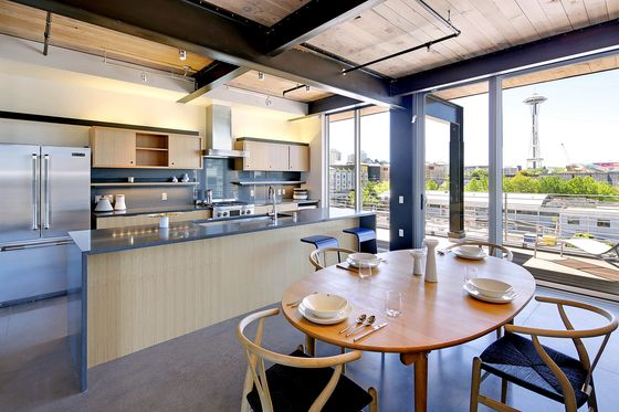 Modern kitchen and dining area with an open floor plan and large floor-to-ceiling windows. The kitchen features stainless steel appliances, light wood cabinetry, and a sleek gray countertop with barstools. A round wooden dining table is set with white dishes and minimal tableware. Outside the windows is a spacious balcony and a clear view of the Space Needle and surrounding cityscape in Seattle.