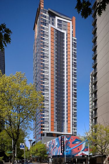 Tall residential skyscraper in downtown Seattle, known as Arrivé, featuring a modern facade with vertical orange and gray panels and expansive windows. At the base of the building is the Cinerama movie theater, with a vibrant, colorful mural on its exterior. The Space Needle is visible in the background, framed by trees and surrounding high-rise buildings on a clear, sunny day.