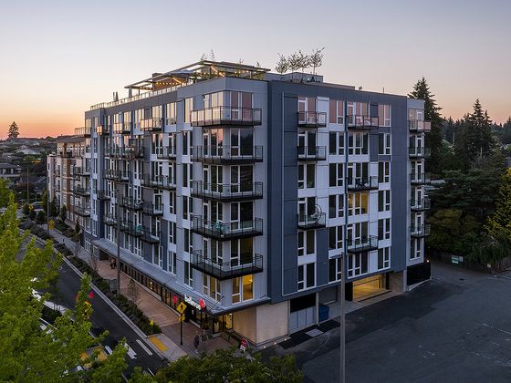 Modern mid-rise apartment building at dusk, featuring large windows, balconies, and a rooftop terrace with string lights. The building is labeled 