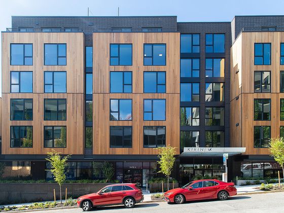 Street view of Kirin Apartments, a modern mid-rise building featuring a mix of natural wood and dark brick exterior, with large grid-style windows and two red cars parked in front.