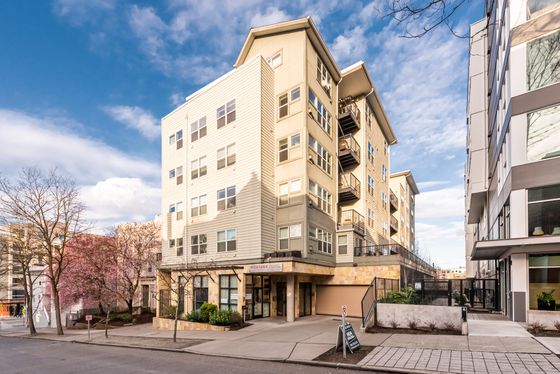 Street view of the Montage apartment building on a sunny day, showing a six-story tan and beige exterior with modern siding, large windows, and small balconies. A sidewalk sandwich board sign is visible near the entrance, with blooming trees and surrounding urban buildings in view.