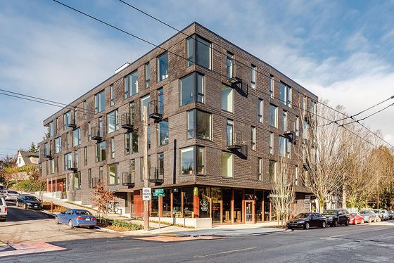 Street view of The Shea, a modern dark brick apartment building with large windows, small balconies, and ground-level retail on a sunny day in a residential neighborhood.