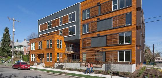 Contemporary apartment building with warm wood siding and dark gray paneling, featuring large windows and a clean, geometric design on a sunny residential street corner.