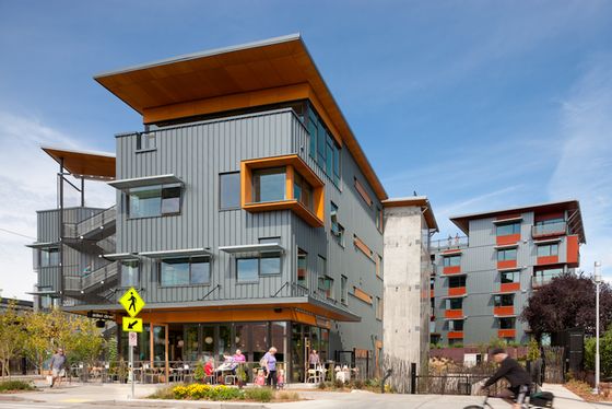 Modern multi-story apartment building with angular rooflines and metal siding, featuring orange window frames and a ground-level restaurant with outdoor seating.