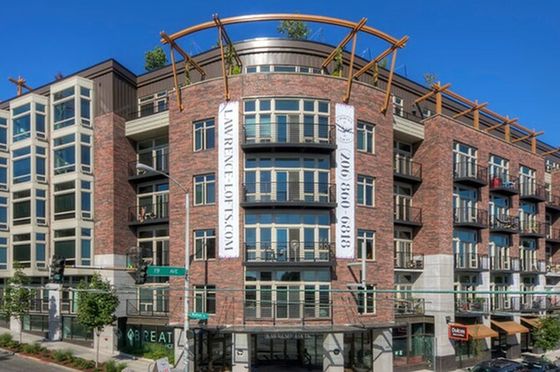 Exterior view of Lawrence Lofts, a modern brick apartment building with large windows, private balconies, and rooftop architectural features, located on a city corner with visible retail on the ground floor.