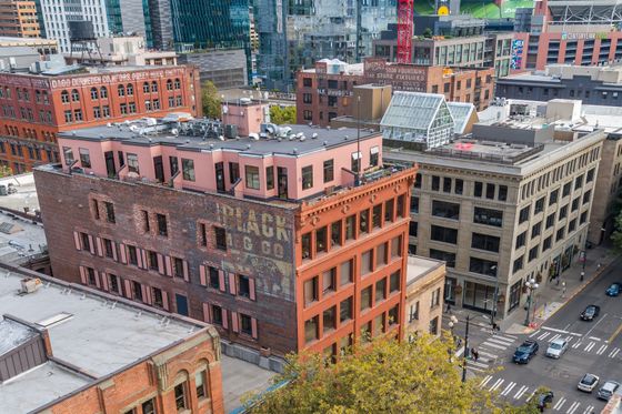 Aerial view of the historic Quilt Building in downtown Seattle, featuring red brick architecture, upper-level pink accents, and faded painted signage, surrounded by modern high-rises and nearby intersections.