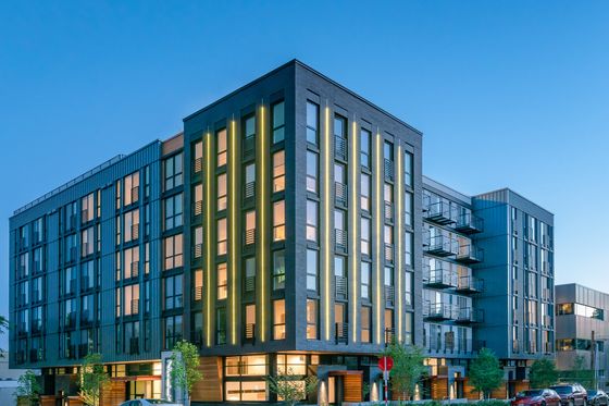 Modern mid-rise apartment building at dusk with illuminated windows and vertical accent lighting, located on a quiet street with parked cars