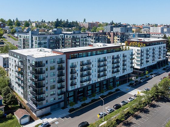 A modern mid-rise apartment building with white and gray exterior, balconies, and rooftop greenery on a sunny day.