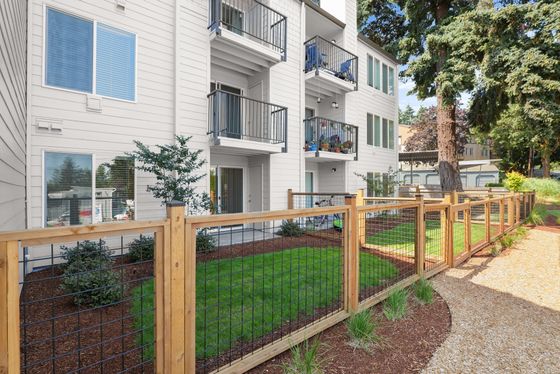 Exterior of The Finch apartments in Vancouver, WA, with beige siding, private balconies, and tall evergreen trees nearby.