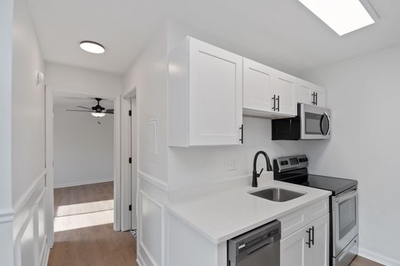 Modern kitchen with sleek white cabinets, stainless steel appliances, and elegant black fixtures, leading to a bright hallway and spacious room.