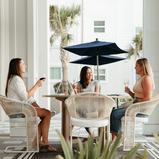 Three people enjoying wine on a stylish terrace with wicker chairs and a blue umbrella.