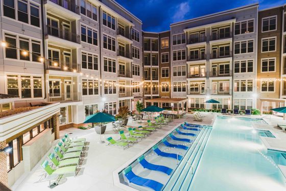 Luxurious apartment complex featuring a modern swimming pool and lounging area illuminated at night, surrounded by stylish multi-story buildings and vibrant patio seating.