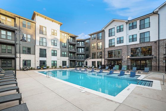 Modern apartment complex featuring a stunning outdoor pool with ample lounge seating, surrounded by stylish multi-story buildings against a clear blue sky.