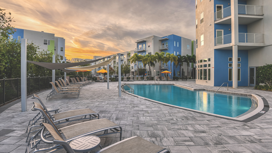 Luxurious apartment complex featuring a sparkling blue swimming pool surrounded by relaxing lounge chairs, vibrant umbrellas, and modern architecture against a stunning sunset backdrop.