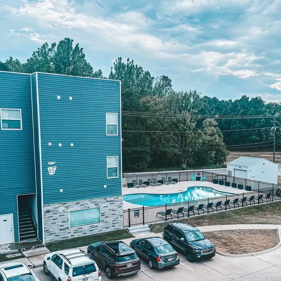 Modern apartment building with vibrant blue exterior, featuring a pristine swimming pool and ample parking surrounded by lush greenery.
