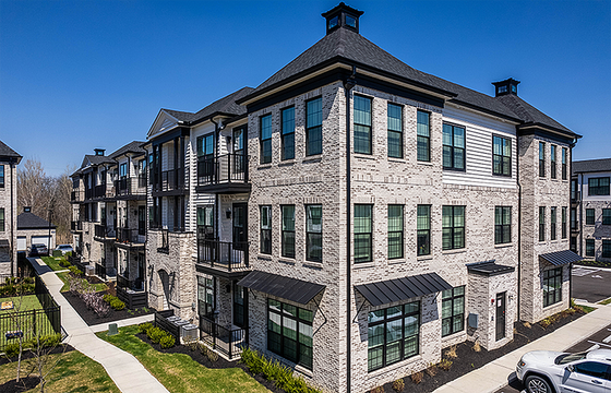 Modern three-story apartment building with elegant brick facade, large windows, and private balconies, set in a landscaped environment on a sunny day.
