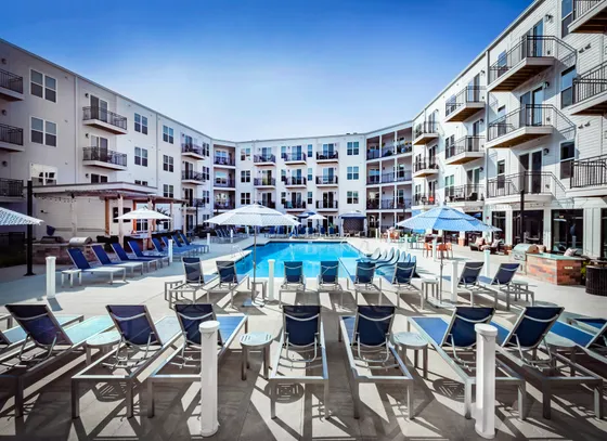 Luxurious apartment complex with a vibrant central pool area, surrounded by modern balconies and relaxing sun loungers under chic umbrella canopies.