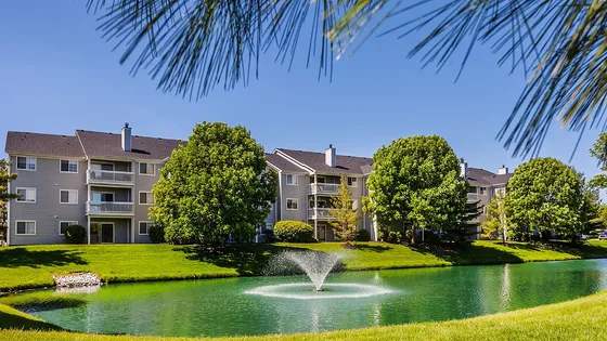 Charming apartment complex with balconies, surrounded by lush greenery and a serene pond with a fountain, offering a picturesque outdoor experience.