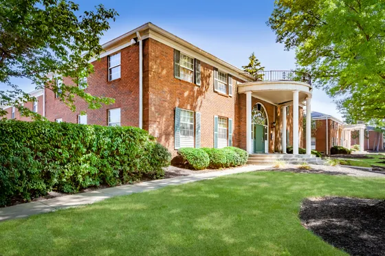Charming brick apartment with elegant columns and lush greenery, offering a welcoming entrance and serene surroundings.