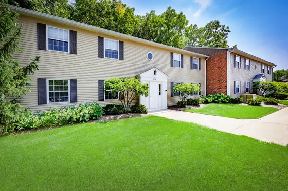 Charming two-story apartment building with beige siding and brick, surrounded by lush green landscaping and vibrant shrubbery.