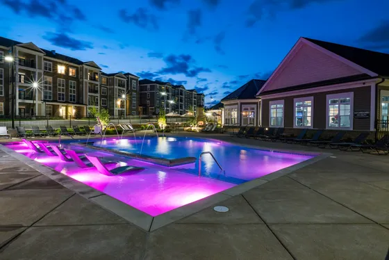 Luxurious apartment complex with illuminated swimming pool featuring elegant lounge chairs and beautifully lit surroundings at dusk.