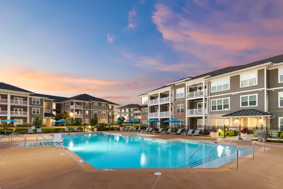 Luxurious apartment complex with a sparkling pool surrounded by elegant buildings and vibrant landscaping under a stunning sunset sky.