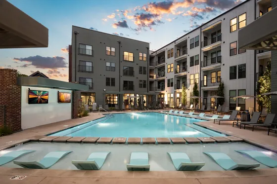 Luxurious apartment complex with a stunning outdoor pool, elegant lounge chairs, and modern architecture under a picturesque sunset sky.