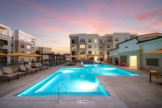 Luxurious apartment complex with a stunning pool area, surrounded by modern buildings and illuminated by a beautiful sunset sky.