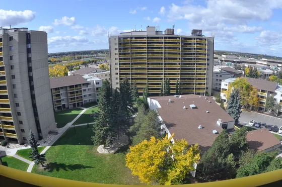 Panoramic view of modern apartment buildings surrounded by lush greenery, showcasing spacious balconies and vibrant fall foliage under a blue sky.