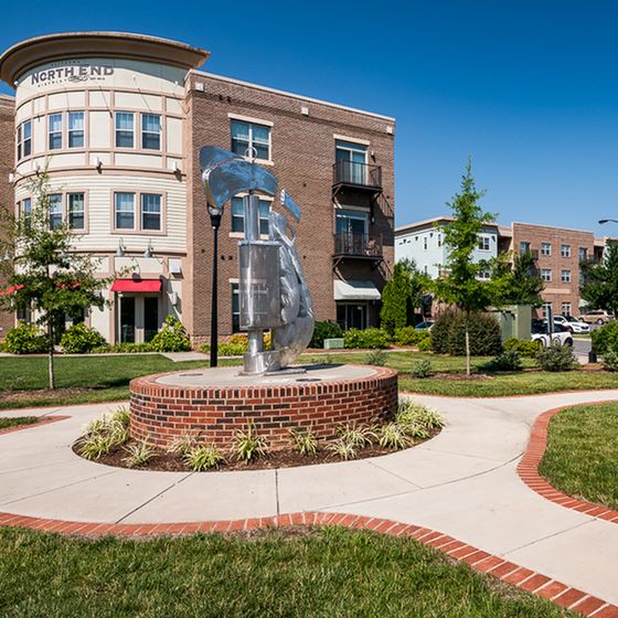 Modern apartment complex with brick and cream exterior, featuring landscaped walkways and a striking silver sculpture centerpiece.