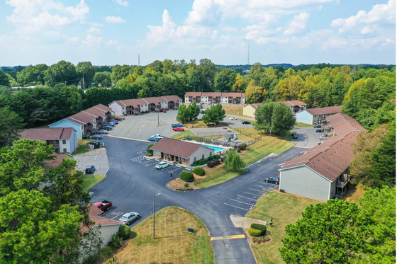 Inviting aerial view of a serene apartment complex nestled in lush greenery, with ample parking and modern buildings.