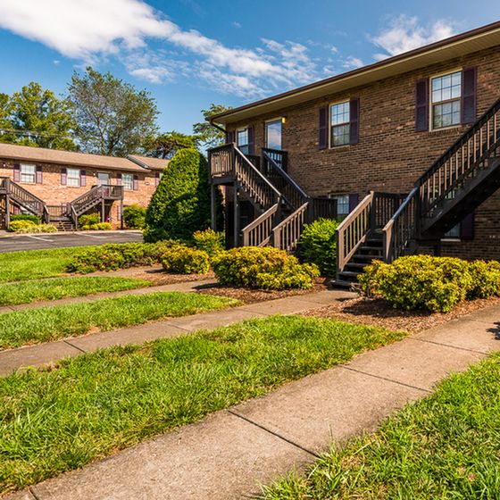 Charming brick apartment building with lush greenery, external staircases, and a vibrant lawn under a clear blue sky.