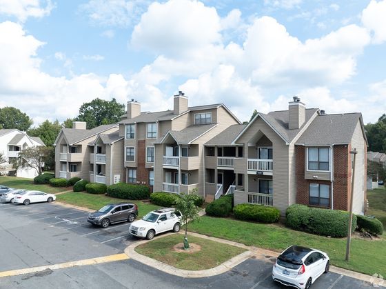 Charming apartment complex with modern architecture, featuring balconies and ample parking, nestled in a lush green setting under a bright blue sky.