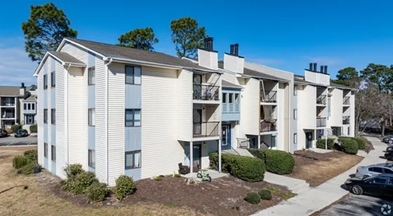 Charming apartment complex exterior with multiple balconies, nestled in a serene setting with lush greenery and ample parking space.