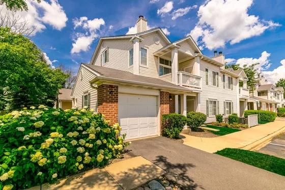 Charming duplex townhouse with a garage, surrounded by lush greenery and vibrant hydrangeas under a clear blue sky.